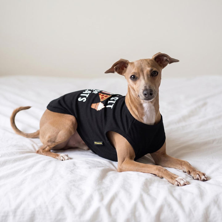Light brown Italian Greyhound dog lying on a white bed, wearing a black sleeveless dog tank top with a white and orange treat print