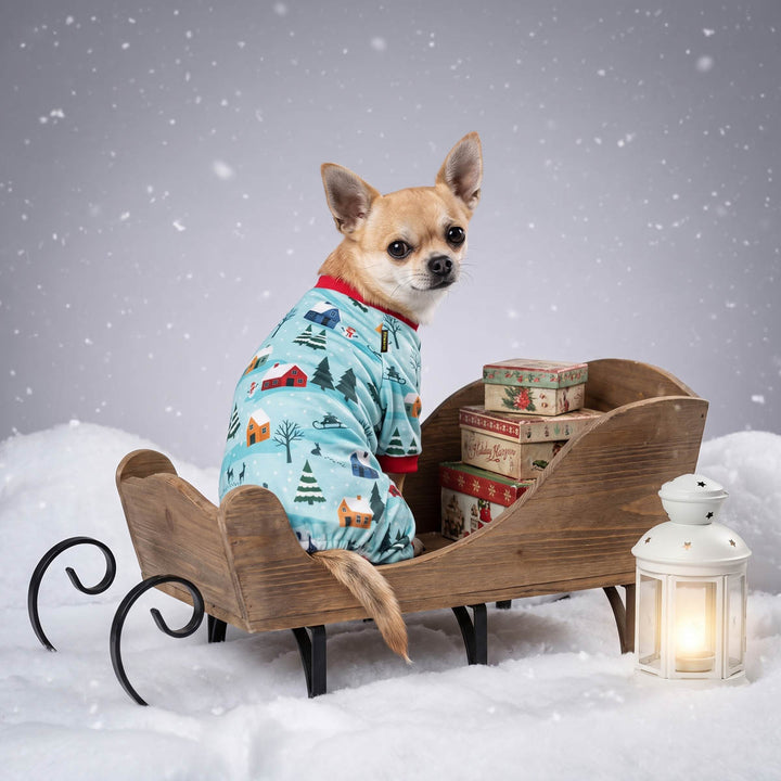Small Chihuahua sits in a wooden sleigh filled with gift boxes, wearing a light blue dog Christmas pajamas with a winter village print