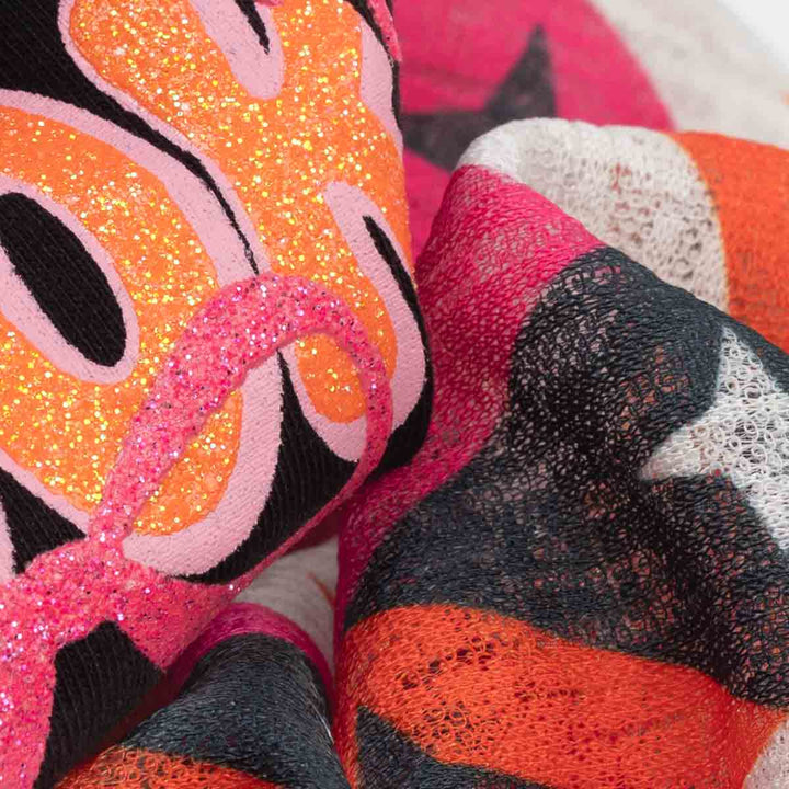 Close-up of a multicolor knit fabric with a bold pattern of orange, pink, black, and white, featuring both glittery and open-knit textures