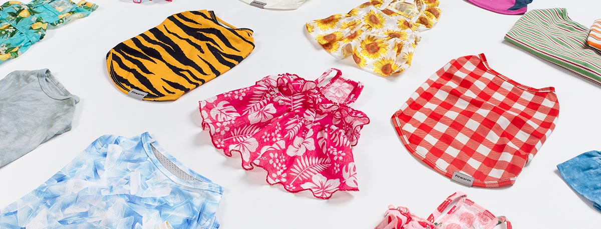 Variety of summer dog clothes spread out, featuring designs like tiger stripes, tropical pink, sunflowers, gingham checks, and tie-dye.
