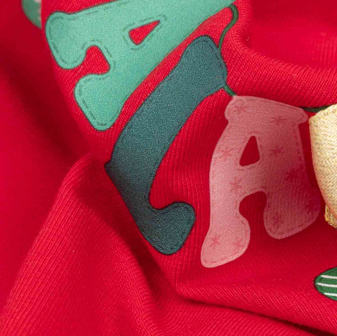 Close-up of a red fabric featuring large decorative letters in mint green, dark teal, and pale pink, with a small gold ribbon bow on the right