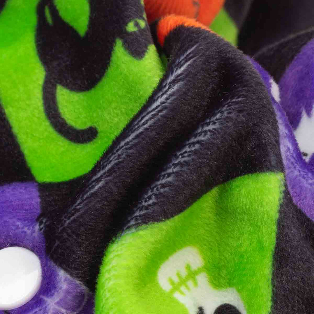 Close-up of a plush fleece dog pjs with a Halloween design, featuring bright green sections with black cat and a white stitched skull, alongside purple and orange sections on a black background