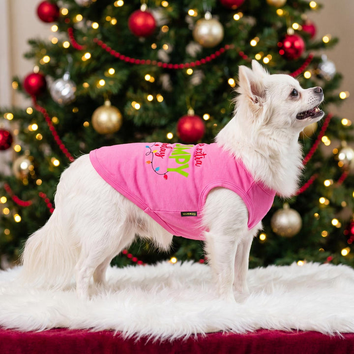 Small white dog stands on a white faux-fur rug in front of a decorated Christmas tree, wearing a bright pink dog tank top with colorful holiday text
