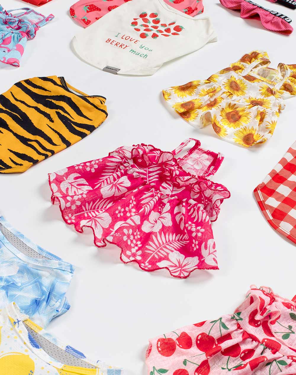 Variety of summer dog clothes spread out, featuring designs like tiger stripes, tropical pink, sunflowers, gingham checks, and tie-dye.