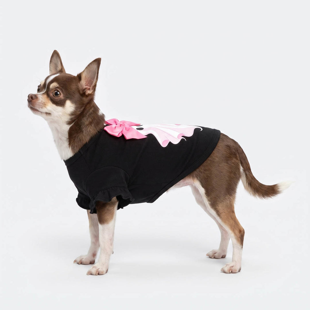 Small brown and white Chihuahua dog wearing a black dog tshirt with a pink bow and pink wing design on the back, standing against a plain light gray studio background