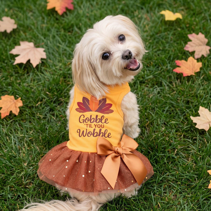 Small white dog sitting on green grass outdoors, wearing a dog Thanksgiving dress with a leaf graphic and the lettering 'Gobble 'til You Wobble,' paired with a brown tutu skirt and large bow