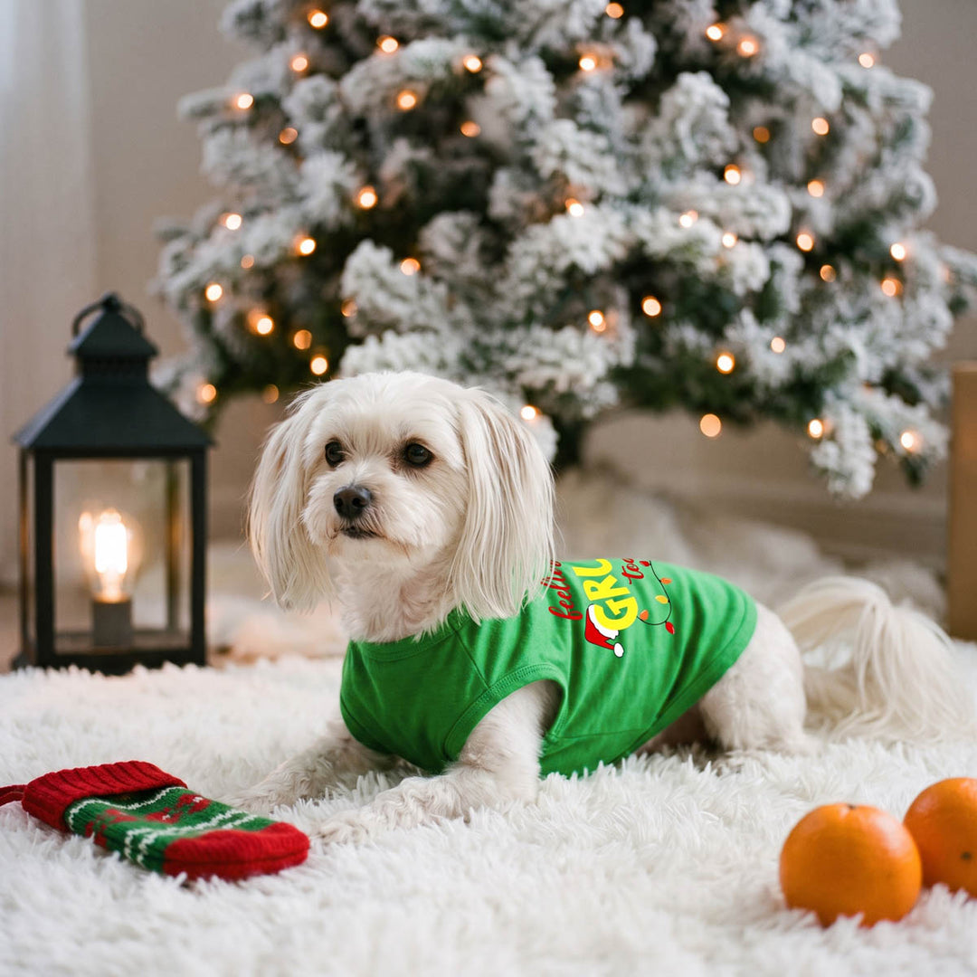 Small white dog wearing a bright green Christmas-themed dog t-shirt lies on a white blanket in front of a decorated Christmas tree