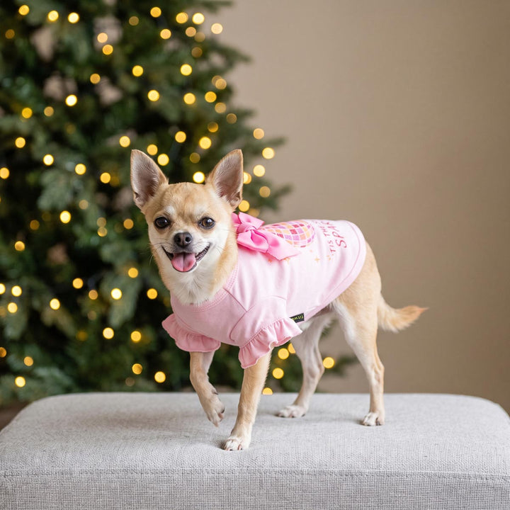 Small tan Chihuahua dog wearing a pink ruffled dog tshirt with a bow and holiday graphics, standing on a gray upholstered bench in front of a blurred Christmas tree with warm lights