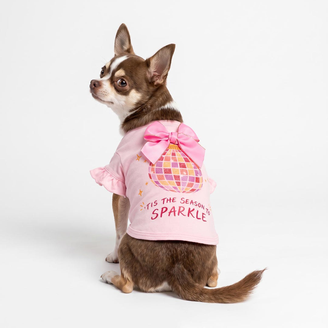 Small brown and white dog wearing a pink ruffled shirt for dogs with a large pink bow, disco ball and 'Tis the season to sparkle', sitting against a plain white studio background