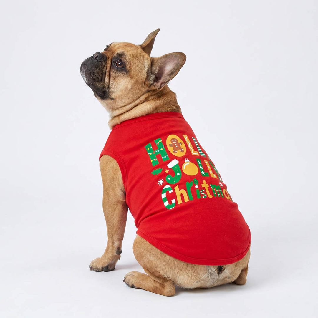 French Bulldog wearing a red sleeveless Christmas dog t-shirt with 'Holly Jolly Christmas' printed in green, white, and gold, sitting against a white studio background