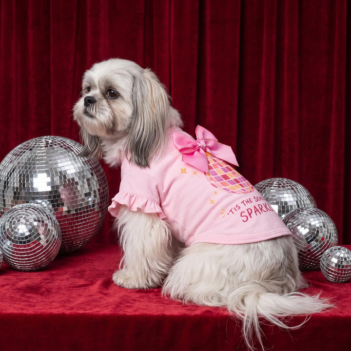 Small fluffy dog sits on a red velvet surface wearing a pink ruffled dog tshirt with a bow and the words 'Tis the season to sparkle