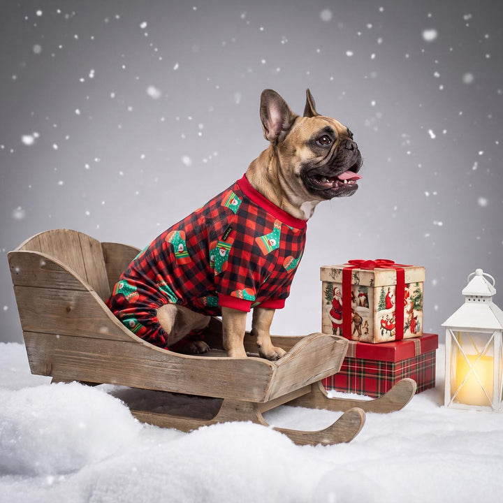 French bulldog wearing red and black plaid Christmas dog pjs sits in a wooden sled on a snowy surface, with wrapped gift boxes and a glowing lantern against a gray background with falling snow