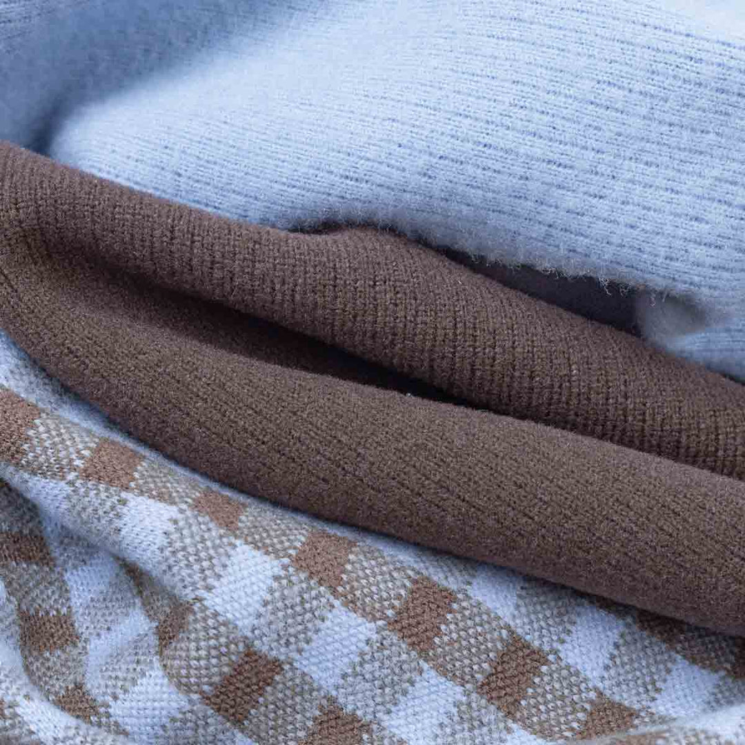 Close-up of folded knit fabric of dog sweaters in three colorways: a light icy blue ribbed knit, a medium brown ribbed knit, and a white and tan checkered knit
