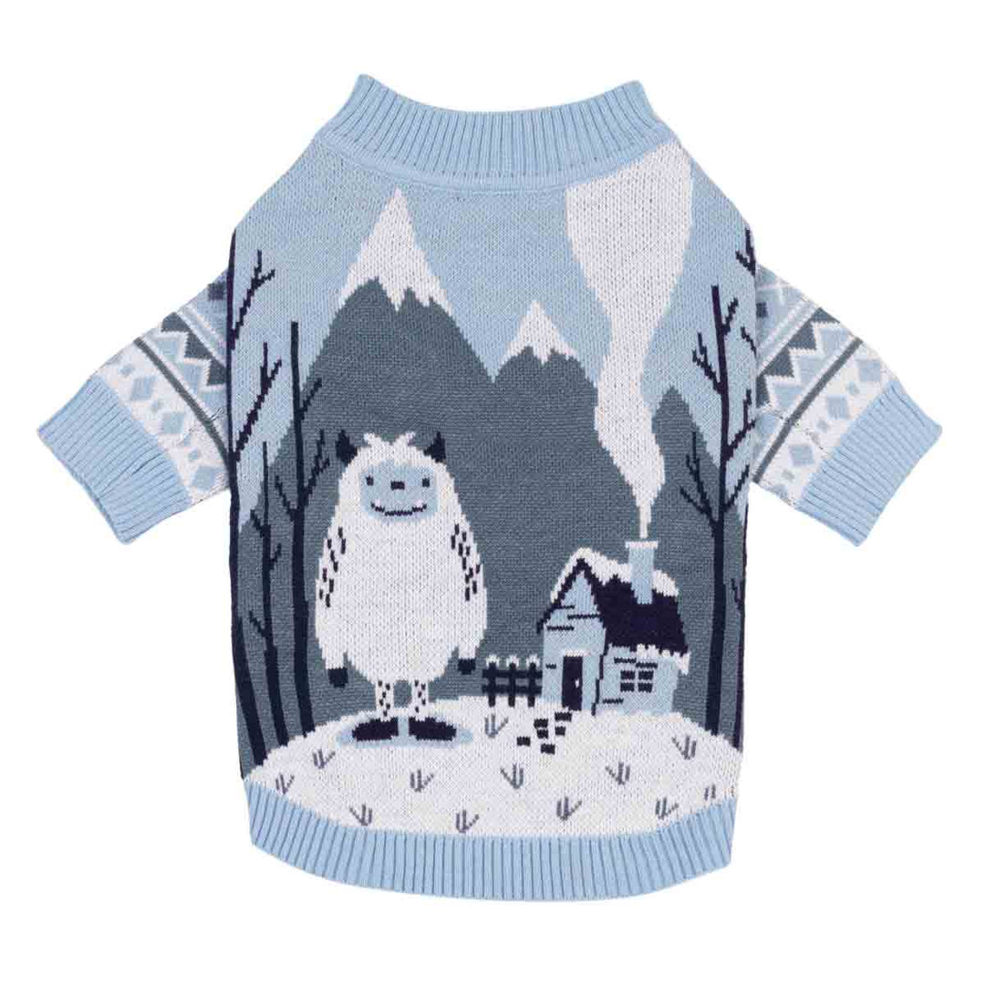 Light blue knitted sweater for dogs featuring a winter mountain scene with a white yeti, a small snowy house, and dark gray trees, shown on a plain white background