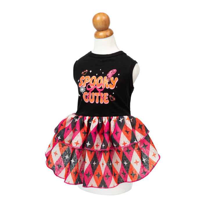 Halloween dog dress displayed on a white mannequin with a wooden neck cap. The dress features a black sleeveless top with 'Spooky Cutie' printed in orange and pink, paired with a tiered ruffle skirt in a black, pink, orange, and white harlequin pattern