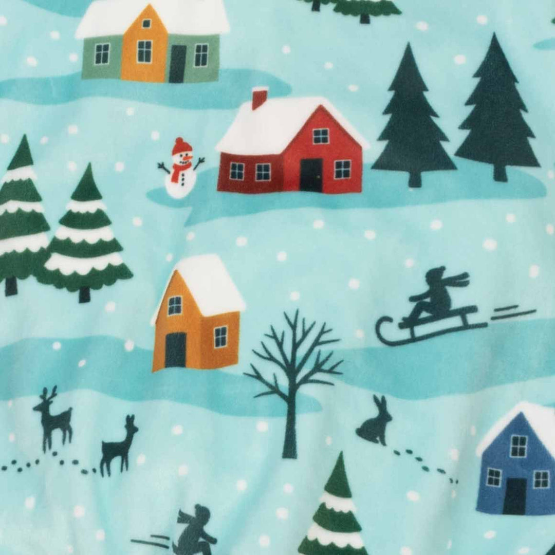 Close-up of a light blue fabric with a winter village print featuring red, orange, and blue houses, dark green pine trees, a snowman, reindeer, rabbits, and sledders