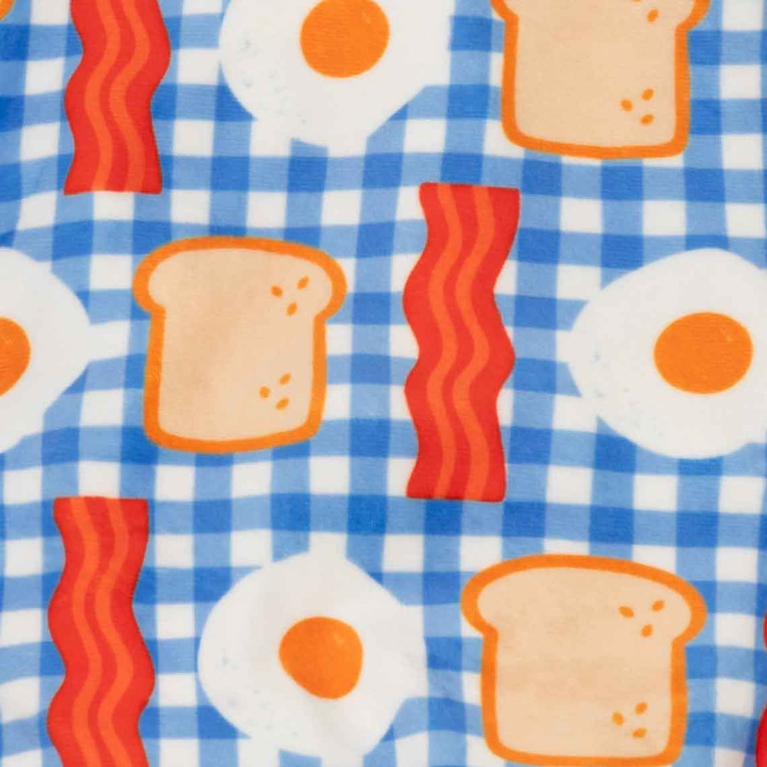 Close-up of a fabric pattern featuring a blue and white gingham background with repeating prints of bacon strips, fried eggs, and slices of toast