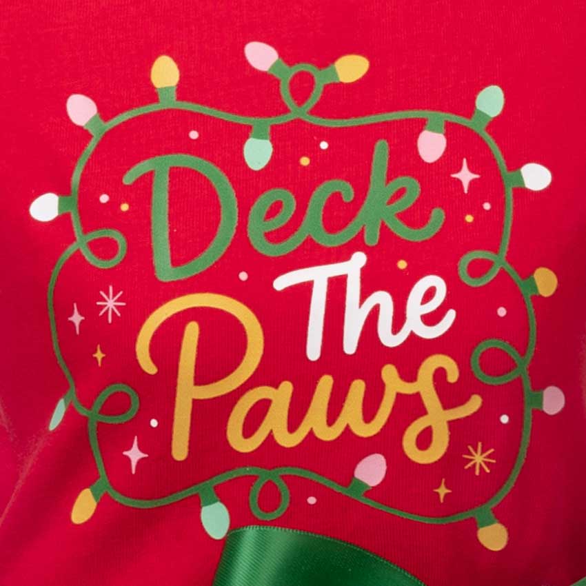 Close-up of a red fabric surface with a decorative holiday graphic reading “Deck The Paws” in green, white, and yellow lettering, surrounded by green string lights and small star accents