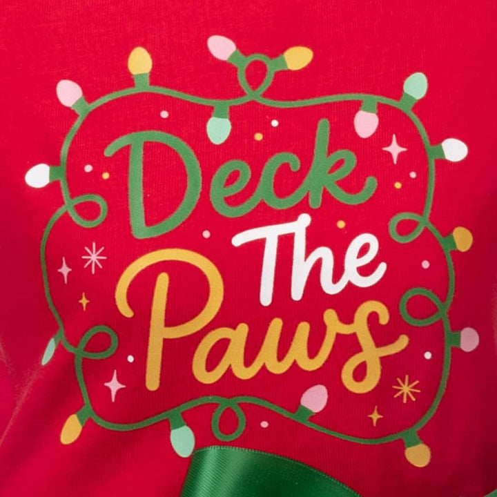 Close-up of a red fabric surface with a decorative holiday graphic reading “Deck The Paws” in green, white, and yellow lettering, surrounded by green string lights and small star accents
