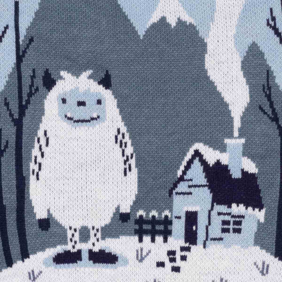 Close-up of a knitted textile featuring a cozy winter scene with a large white yeti-like character, a small house with a smoking chimney, dark trees, and snowy ground in muted blue-gray, white, and navy tones