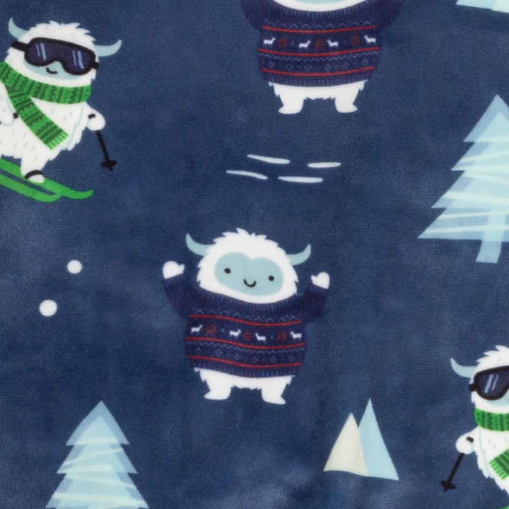 Close-up of a navy blue fabric with a repeating winter pattern featuring cartoon white yetis in colorful sweaters, light blue Christmas trees, and small white snowflakes