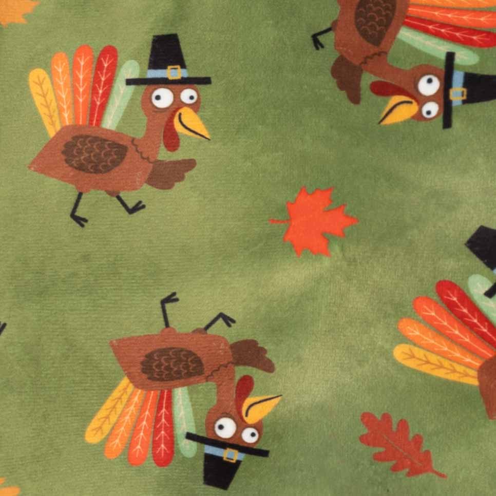 Close-up of fabric of Thanksgiving dog onesies featuring a repeating pattern of cartoon turkeys wearing black pilgrim hats and colorful autumn leaves on an olive green background