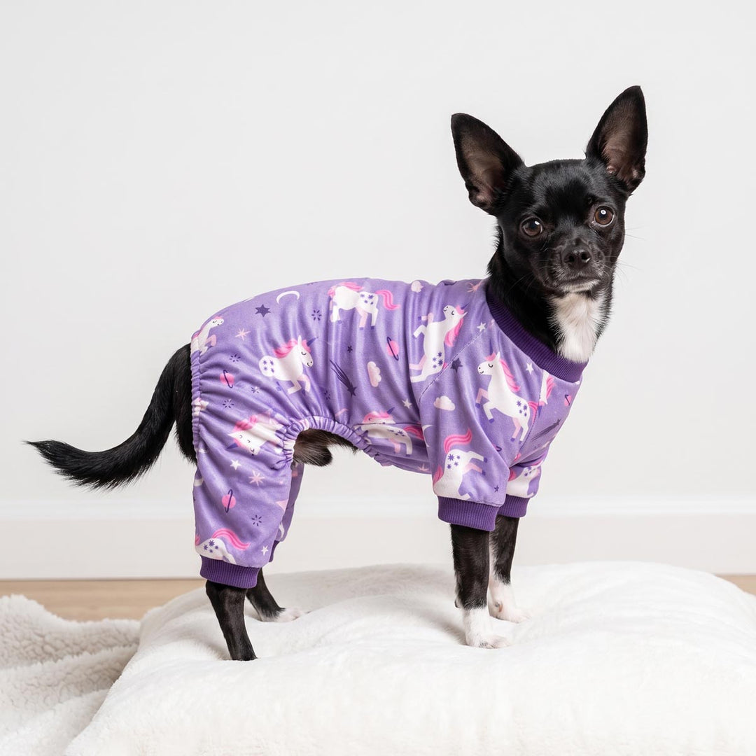Small black and white dog wearing a purple full-body dog onesie with a unicorn print, standing on a white cushion against a plain light background