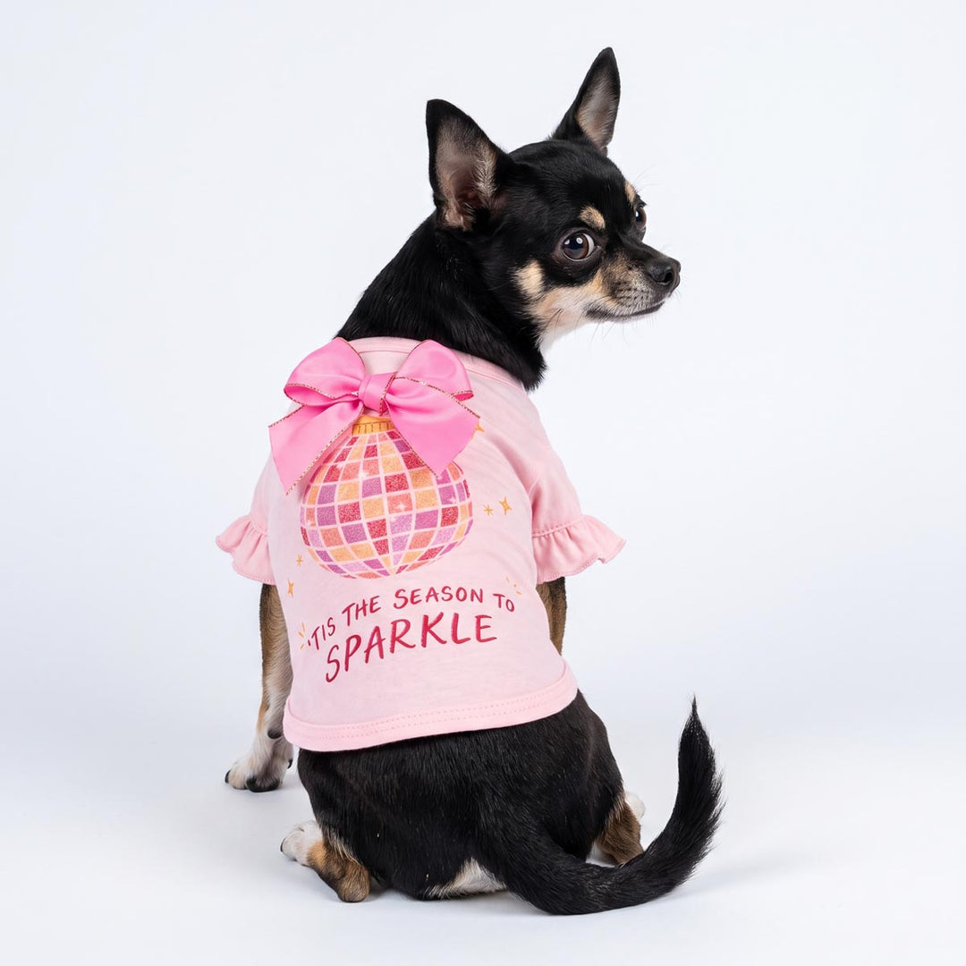 Small black and tan dog wearing a pink ruffled dog tshirt with a large pink bow and a disco ball graphic, sitting against a white studio background