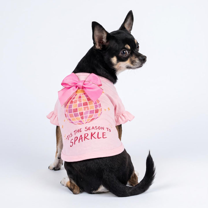 Small black and tan dog wearing a pink ruffled dog tshirt with a large pink bow and a disco ball graphic, sitting against a white studio background